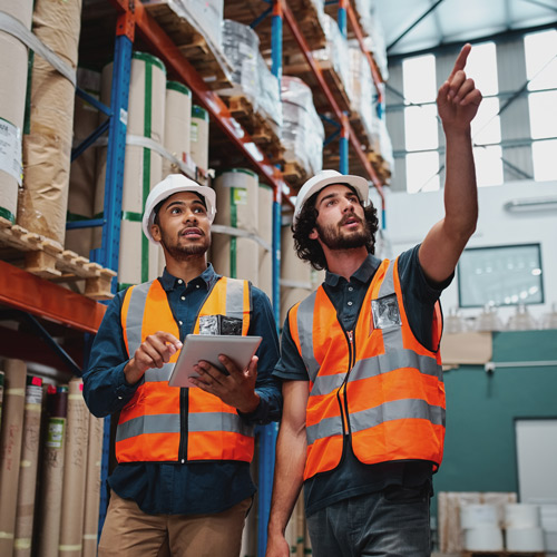 Two warehouse workers in safety vests and helmets examining shelves, one holding a tablet and the other pointing upward.